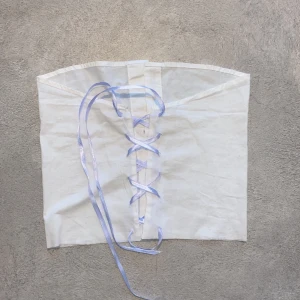 Vit topp med blå snörning - Unik vit korsett-tubtopp i skjortmaterial med snygg snörning baktill i ljusblått satinband och knappar framtill. Toppen har en cool skjortkrage som kan stylas på olika sätt för en personlig look. Perfekt för dig som vill sticka ut med en kreativ och edgy vibe.