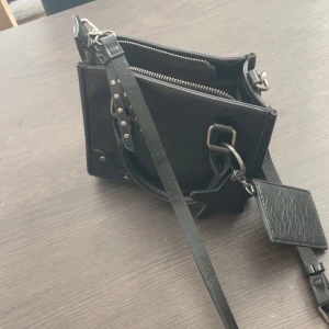Svart axelväska med silverdetaljer - Snygg svart axelväska i skinn med justerbar axelrem och coola silverdetaljer. Väskan har flera fack med dragkedja och en dekorativ tag på sidan. Perfekt för att bära dina viktigaste grejer och ge outfiten en edgy touch.