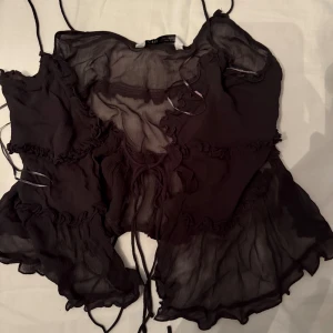 Svart linnetopp från Zara - Supersöt svart linnetopp från Zara med tunna axelband och volangdetaljer. Toppen är gjord i ett lätt, transparent tyg och har snörning framtill. Perfekt för dig som gillar en romantisk och trendig look.