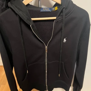 Svart hoodie från Polo Ralph Lauren - Svart hoodie från Polo Ralph Lauren med dragkedja och huva. Tröjan har ett broderat vitt logomärke på bröstet, dragsko vid huvan och mjukt foder på insidan. Perfekt för kyliga dagar och enkel att matcha med andra plagg. Storlek S. Tröjan är knappt använd bara en skoldag, inga defekter eller liknanden, priset kan diskuteras vid snabb affär.