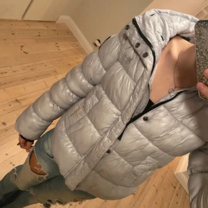 Silver pufferjacka från Superdry XP-D - Säljer en riktigt snygg silverfärgad pufferjacka från Superdry XP-D. Jackan har en glansig finish, vadderad design och huva. Detaljer som broderad logga på bröstet, svart patch på ärmen och dragkedja framtill. Perfekt för kalla dagar när du vill sticka ut.