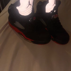  Nike Air Jordan 5 sneakers - Säljer ett par svarta Nike Air Jordan 5 sneakers med röda detaljer och klassisk Jumpman-logga på plösen. Skorna har snörning, platt sula och meshpaneler på sidorna.