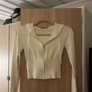 Vit croppad hoodie från Catania - Snygg croppad vit kofta från Catania med dragkedja framtill och ribbade detaljer vid midjan och ärmsluten. Tröjan har långa ärmar och huva med snörning, perfekt för en chill och trendig look.