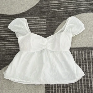 Vit blus med puffärm från Hollister - Supersöt vit blus från Hollister med puffiga korta ärmar och v-ringning. Toppen har en flowy passform och snygga korsade band samt öppen rygg. Perfekt för sommarens alla tillfällen och ger en romantisk vibe. Tillverkad i ett lätt och luftigt material.