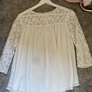 Vit blus med spetsdetaljer - Säljer en vit blus med vackra spetsdetaljer upptill och på ärmarna. Blusen har rund halsringning, trekvartsärmar och knäppning bak i nacken. Spetsen har ett blommigt mönster som ger en romantisk känsla. Perfekt att styla med jeans eller kjol. OPSdet är min mobilkamera om är lite sönder så det är det  som gör att det blir en fläck på bilden.!🥰