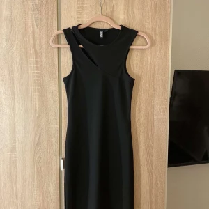 Svart asymmetrisk klänning från Pieces - Säljer en svart, tight och stilren klänning från Pieces i storlek XS. Klänningen har en cool asymmetrisk halsringning och är ärmlös, vilket ger en modern vibe. Perfekt för dig som gillar minimalistisk stil och vill sticka ut lite extra.