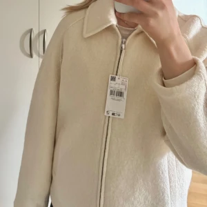 Beige/Krämvit ullhacka med dragkedja - Säljer en stilren ulljacka med krage och dragkedja framtill. Jackan har en mjuk, fluffig yta och raka ärmar. Perfekt för dig som gillar en clean look och vill ha något som är både snyggt och bekvämt till hösten/våren. Helt ny, med prislappen på. Storlek S.  Rek pris 899kr på Mango. 