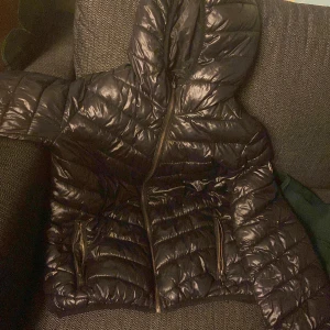 Svart pufferjacka från FB Sister - Svart glansig pufferjacka från FB Sister med huva och dragkedja framtill. Jackan har två fickor med dragkedja och quiltad design. Perfekt för kyliga dagar och har en sportig vibe.