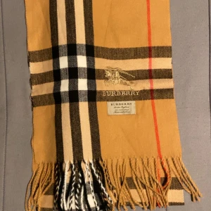 Rutig halsduk från Burberry i ull - Klassisk halsduk från Burberry med ikoniskt rutigt mönster i beige, svart, vitt och röda detaljer. Halsduken har fransar i ändarna och broderad Burberry-logga. Perfekt accessoar för att lyfta din outfit med en tidlös brittisk touch.