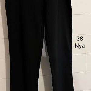 Svarta kostymbyxor med dragkedja - Svarta kostymbyxor med rak passform och diskret dragkedja i sidan. Byxorna har en klassisk look och är tillverkade i ett slätt tyg som ger en stilren känsla. Perfekta för dig som gillar en clean och enkel stil.