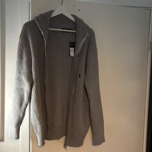 Grå stickad hoodie från Polo Ralph Lauren - Säljer en grå stickad hoodie från Polo Ralph Lauren i storlek XL. Tröjan har huva, dragkedja och ribbad struktur. Klassisk logga broderad på bröstet och riktigt skön passform. Perfekt för dig som gillar stilren och avslappnad look.