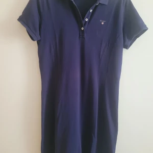 Marinblå pikéklänning från Gant - Snygg marinblå pikéklänning från Gant med klassisk krage och knappar framtill. Klänningen har korta ärmar och är tillverkad i mjuk bomull. Perfekt för en avslappnad och sportig look med broderad Gant-logga på bröstet.