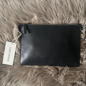 Svart kuvertväska från Carin Wester  - Stilren svart kuvertväska från Carin Wester. Väskan har minimalistisk design med dragkedja upptill och diskret logga framtill. Oanvänd med prislapp kvar. Långt band följer även med.