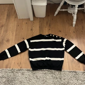 Randig svartvit tröja från H&M XS - Svart tröja från H&M i storlek XS med breda vita ränder och snygga guldknappar på ena axeln. Tröjan har lång ärm och ribbade muddar vid hals, ärmslut och nederkant. Perfekt för dig som gillar en klassisk men trendig look. Aldrig använt den.