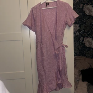 Ljuslila omlottklänning Vero Moda - Superfin ljuslila omlottklänning från Vero Moda med kort ärm, volangkant nertill och knyt i midjan. Klänningen har ett diskret prickigt mönster 