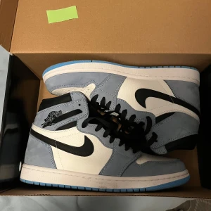 Nike Air Jordan 1 High University Blue - Nike Air Jordan 1 High i färgen University Blue med vit läderbas, ljusblå mockadetaljer och svarta swoosh-loggor. Skorna har svarta snören, hög siluett och klassisk Air Jordan Wings-logga på sidan. Perfekta för dig som vill sticka ut med en ikonisk sneaker.