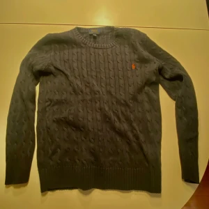 Mörkblå kabelstickad tröja Polo Ralph Lauren - Kabelstickad mörkblå tröja från Polo Ralph Lauren med klassisk rund hals och röd broderad logga på bröstet. Långärmad och ribbad vid ärmslut och nederkant. Perfekt för dig som gillar stilren och tidlös design.