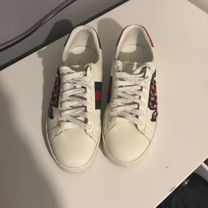 Gucci Ace sneakers med orm brodyr - Säljer ett par vita Gucci Ace sneakers i skinn med ikoniska gröna och röda ränder på sidorna samt broderad orm. Baksidan har detaljer i rött och grönt. Klassisk rund tå och snörning framtill. Perfekta för dig som vill sticka ut med lyxig streetstyle.