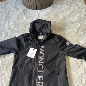 Svart vindjacka från Moncler - Svart vindjacka från Moncler med stor logotyp tryckt längs dragkedjan och klassisk Moncler-patch på ärmen. Jackan har huva, två sidofickor med dragkedja och snygga detaljer. Perfekt för dig som gillar stilrena och sportiga plagg.
