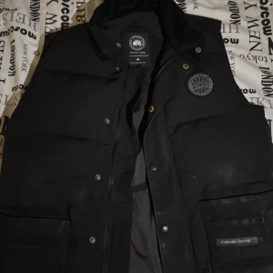 Black label Canada Goose - Svart äkta Canada Goose väst med black label med klassisk logga på bröstet och diskreta knappar framtill. Västen har två stora fickor med lock och dragkedja samt hög krage. Perfekt för lager-på-lager och riktigt snygg till streetwear.