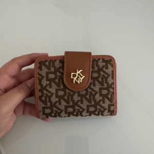 Brun DKNY plånbok med monogram - Snygg plånbok från DKNY i brunt med klassiskt monogrammönster och guldfärgad logga. Plånboken har flera kortfack, fönster för ID och dragkedjeförsett myntfack. Tillverkad i skinn och textil med stilrena detaljer.