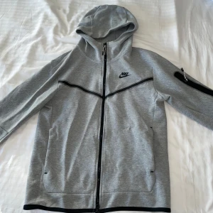 Grå Nike hoodie med dragkedja - Snygg grå hoodie från Nike med hel dragkedja och svarta detaljer. Tröjan har huva, långa ärmar och en praktisk ficka med dragkedja på ena ärmen. Perfekt för chill eller träning, klassisk logga på bröstet.