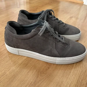 Grå mocka sneakers från Axel Arigato - Snygga grå sneakers från Axel Arigato i mocka med vita, platta gummisulor och klassisk snörning. Stilren design med diskret logga på sidan och låg profil. Perfekta för dig som gillar minimalistisk och modern stil.