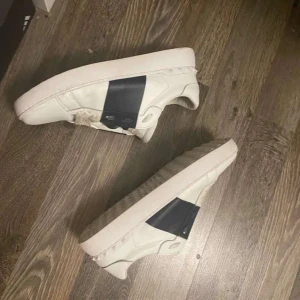 Valentino Garavani vita sneakers - Snygga vita sneakers från Valentino Garavani med bred svart detalj över sidan. Skorna är i skinn och har klassisk snörning samt platt sula. Kommer med originalkartong, dustbag och äkthetsbevis. Perfekta för dig som vill ha en clean och lyxig look.