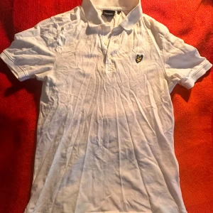 Vit Lyle & Scott pikétröja i bomull - Vit pikétröja från Lyle & Scott med klassisk krage och knappslå framtill. Tröjan har korta ärmar och den ikoniska gulsvarta logon broderad på bröstet. Tillverkad i mjuk bomull, perfekt för en clean och enkel stil.