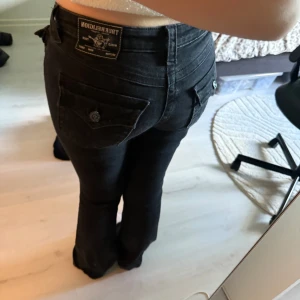 Svarta bootcut jeans från True Religion - Säljer ett par svarta jeans från True Religion med bootcut passform. Byxorna har snygga detaljer som stora fickor med lock och knapp bak, samt dekorativa sömmar. Klassisk jeanslook  och coola knappar med logga. Säljer dom för dom inte kommer till användning längre. Dom heter Becca W26 är storlek ungefär XS/S