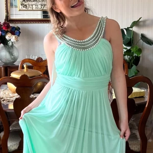 Mintgrön långklänning med pärldetaljer - Superfin mintgrön långklänning med plisserad kjol och bred midja. Klänningen har smala axelband och en snygg pärlkrage som ger extra wow-känsla. Tyget är lätt och faller snyggt, perfekt för dig som vill sticka ut med en elegant look. Lappar kvar och aldrig använd endast provad 