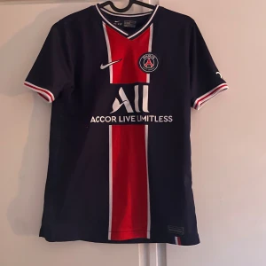 PSG matchtröja Nike Dri-Fit  - Paris Saint-Germain fotbollströja från Nike i Dri-Fit-material. Mörkblå med röd vertikal rand och vita detaljer, PSG-emblem på bröstet och sponsortryck. V-ringad krage med röd, vit och blå kant. Korta ärmar med randiga muddar.