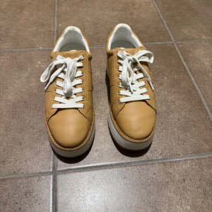 Beige sneakers från Lauren Ralph Lauren - Snygga beige sneakers från Lauren Ralph Lauren med RL-logga på sidan. Skorna har vit platt sula, vita snören och är gjorda i skinn. Insidan är fodrad i vitt tyg och detaljerna har dekorativa sömmar. Perfekta för dig som gillar stilrena och trendiga sneakers. Använda 1 gång helt nya.