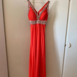 Röd/orange långklänning med paljettdetaljer - Snygg röd/orangea långklänning med djup v-ringning och breda axelband. Klänningen har glittriga paljettdetaljer längs ringningen och midjan, samt plisserad överdel. Perfekt för bal eller festliga tillfällen.