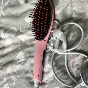 Elektrisk värmeborste - Rosa elektrisk värmeborste för hårstyling. Borsten har svarta piggar med orange toppar och en lång vit sladd. Synligt i gott skick utan tydliga repor eller skador. Perfekt för att snabbt och enkelt styla håret hemma.