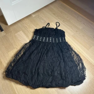 Svart spetsklänning från WeTweens - Supersöt svart kortklänning till barn från WeTweens med tunna axelband och spets över hela klänningen. Midjan har ett brett band med silvriga nitar som ger extra edge. Perfekt för dig som gillar en cool och feminin stil