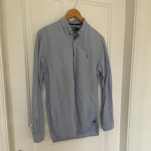 Ljusblå skjorta från Polo Ralph Lauren - Klassisk ljusblå skjorta från Polo Ralph Lauren i slim fit-modell. Skjortan har button down-krage, broderad logga på bröstet och vita knappar. Tillverkad i mjuk bomull och har långa ärmar. Perfekt för en clean och stilren look.
