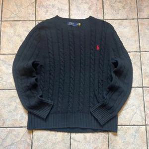 Svart kabelstickad tröja Polo Ralph Lauren - Svart kabelstickad tröja från Polo Ralph Lauren med klassisk röd logga broderad på bröstet. Tröjan har rund hals, ribbade muddar och är långärmad. Perfekt för dig som gillar stilren och tidlös design 📏Måtten på tröjan är 63 cm på längden, 47 cm på bredden, 59 cm på ärmlängden och sitter på size S/170 👌Tröjan är i 9/10 skick, nästan aldrig använd, inga tecken på defekter. Pris går såklart att diskutera 🙌