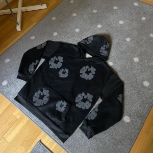 Denim Tears Hooide svart/grå - Svart hoodie från Denim Tears med unika gråa blommönster tryckta över hela tröjan. Klassisk känguruficka framtill och huva. Tillverkad i mjuk bomull, perfekt för dig som vill sticka ut med en streetwear vibe. 