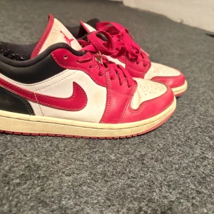 Nike Air Jordan 1 Low röd/vit - Säljer ett par Nike Air Jordan 1 Low i klassisk röd, vit och svart färg. Skorna har röd swoosh, röda snören och detaljer, samt perforerad tåbox. Tillverkade i skinn med platt sula och rund tå. Perfekta för dig som gillar streetstyle och sneakers med ikonisk design.