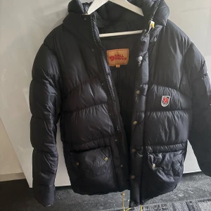 Svart dunjacka från Fjällräven XXS - Säljer en svart, fluffig dunjacka från Fjällräven i storlek XXS. Jackan har huva, dragkedja och tryckknappar framtill, samt två stora fickor med lock. Klassisk Fjällräven-logga på bröstet och insidan. Perfekt för kalla dagar! Aldrig använt då jag söker en annan. Inget fel på den💕Det ser ut som att jackan är blå på sista bilden men det är bara för blixten