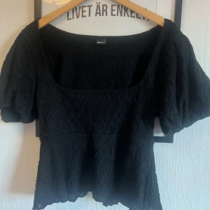 Svart blus med puffärm Gina Tricot - Säljer en svart blus från Gina Tricot i storlek M. Toppen är köpt från Nelly men märket är Gina. Toppen har puffiga korta ärmar, fyrkantig ringning och ett snyggt strukturerat tyg som ger en cool känsla. Passformen är lite stor  så skulle passa en L och materialet känns som polyester. Perfekt för dig som gillar trendiga detaljer. Toppen är endast provad, jag säljer pga att den är för stor för mig i urringningen.   Pris kan diskuteras