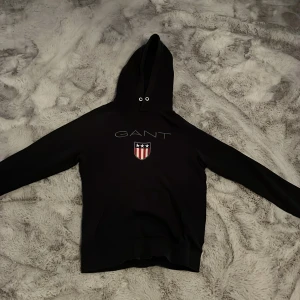 Svart hoodie från GANT med logga - Svart hoodie från GANT med stor logga och sköld på bröstet. Klassisk huva med snörhål och långa ärmar. Perfekt för dig som gillar stilren och sportig look. Mjuk och bekväm, passar till allt.