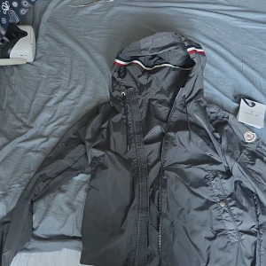 Svart vindjacka från Moncler med huva - Svart vindjacka från Moncler med klassisk logga på ärmen och snygga röd-vit-blå detaljer på huvan. Jackan har dragkedja framtill, justerbar huva med snörning och två fickor med tryckknapp. Perfekt för dig som vill ha en stilren och sportig look.