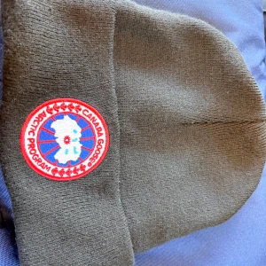 Mörkgrå mössa från Canada Goose - Snygg mörkgrå mössa från Canada Goose med klassisk rund logotyp-patch framtill. Mössan är ribbstickad och tillverkad i 100% akryl, vilket gör den mjuk och skön att bära. Perfekt för kalla dagar och enkel att matcha med din vinterstil.