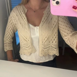 Beige stickad kofta  - Supersöt beige stickad kofta med vackert hålmönster och trekvartsärm. Koftan är croppad och har en öppen design utan knappar, perfekt att slänga över ett linne eller klänning. Ger en romantisk och bohemisk vibe till din outfit.