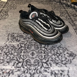 Nike Air Max 97 svart/röd sneakers - Säljer ett par Nike Air Max 97 sneakers i svart med röda detaljer och reflekterande vita ränder. Skorna har den ikoniska vågiga designen, synlig Air-sula och snörning. Perfekta för dig som gillar streetwear och vill sticka ut med en klassisk modell.