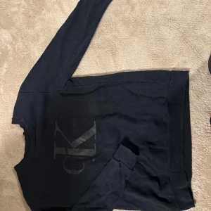 Svart Calvin Klein Jeans sweatshirt - Svart sweatshirt från Calvin Klein Jeans med stor CK-logga framtill. Klassisk rund halsringning och långa ärmar. Mjuk insida och ribbade muddar vid ärmslut och nederkant. Perfekt för en clean och stilren look. Köpte den direkt från NK Gbg för ett par årsen och nu kommer den inte till så stor användning den passar iaf som en 170!