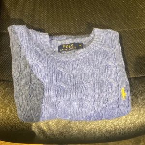 Blå kabelstickad tröja Polo Ralph Lauren - Kabelstickad blå tröja från Polo Ralph Lauren med klassisk gul broderad logga på bröstet. Tröjan har rund halsringning, ribbade muddar och lång ärm. Perfekt för lager-på-lager och snygg till jeans eller kjol. 