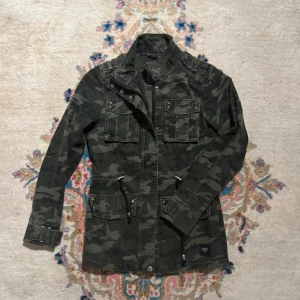 Camouflage fieldjacka - Snygg fieldjacka i camouflage. Jackan har flera fickor med lock och nitar, dragkedja och knappar framtill samt coola axeldetaljer. Storlek S men sitter som XS